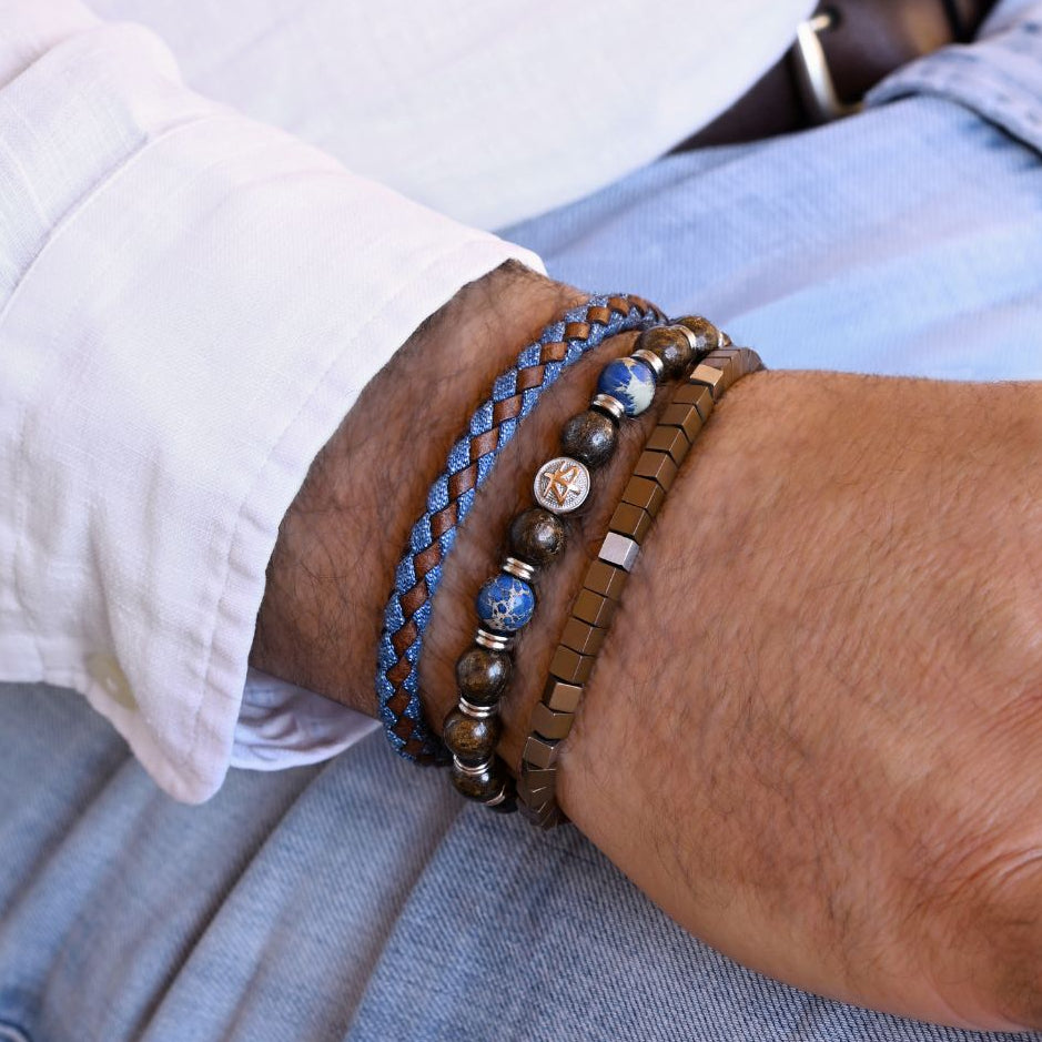 A close-up of a person's wrist wearing multiple bracelets, including a blue braided cord bracelet, a brown beaded bracelet with a central round charm, and a textured brown leather bracelet, paired with a white shirt and light blue jeans.