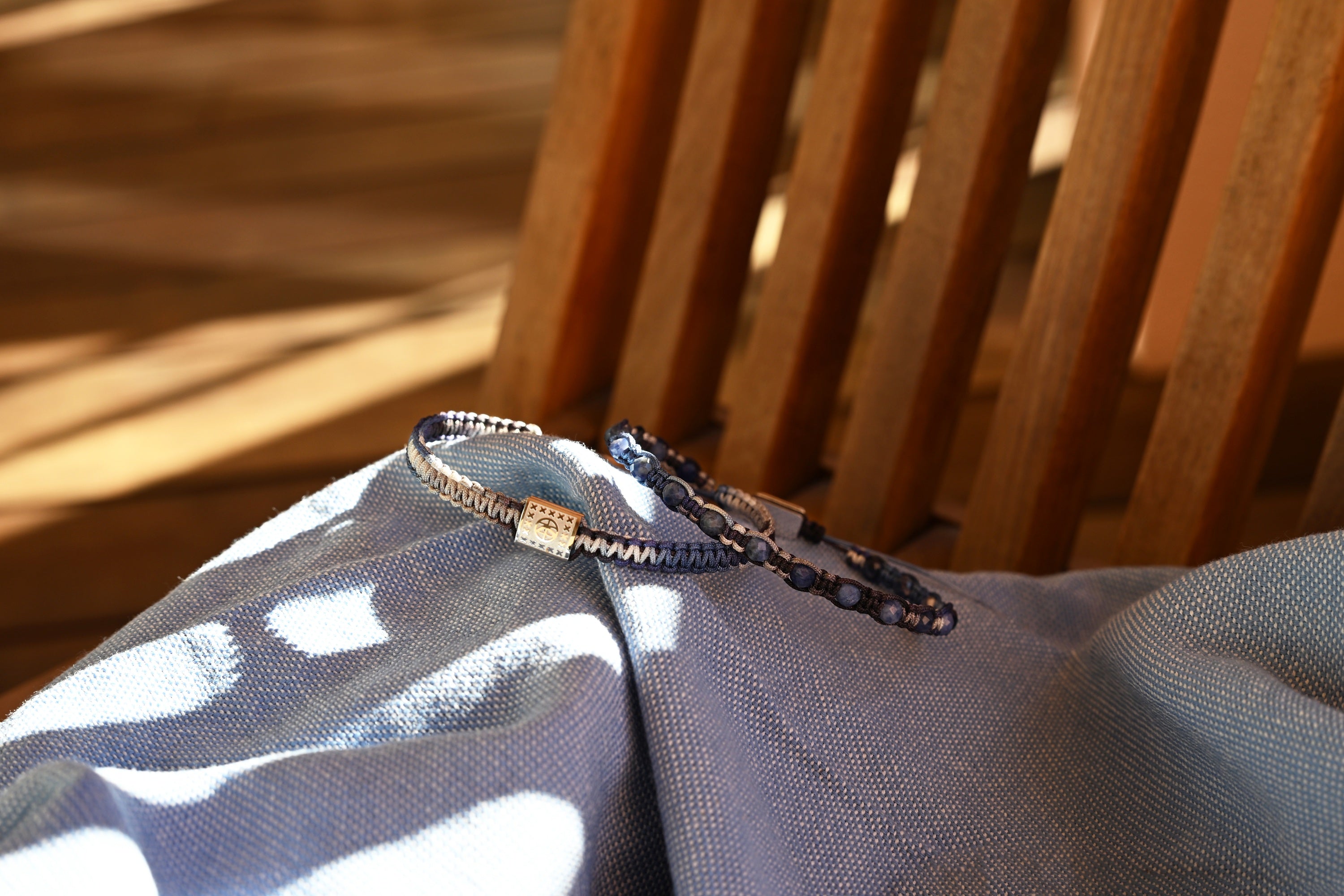 A close-up of two beaded bracelets resting on a light blue fabric surface with a wooden slatted chair in the background. One bracelet is metallic silver centerpiece with gradient blue cord and the other is gradient blue cord with blue lapis lazuli beads.