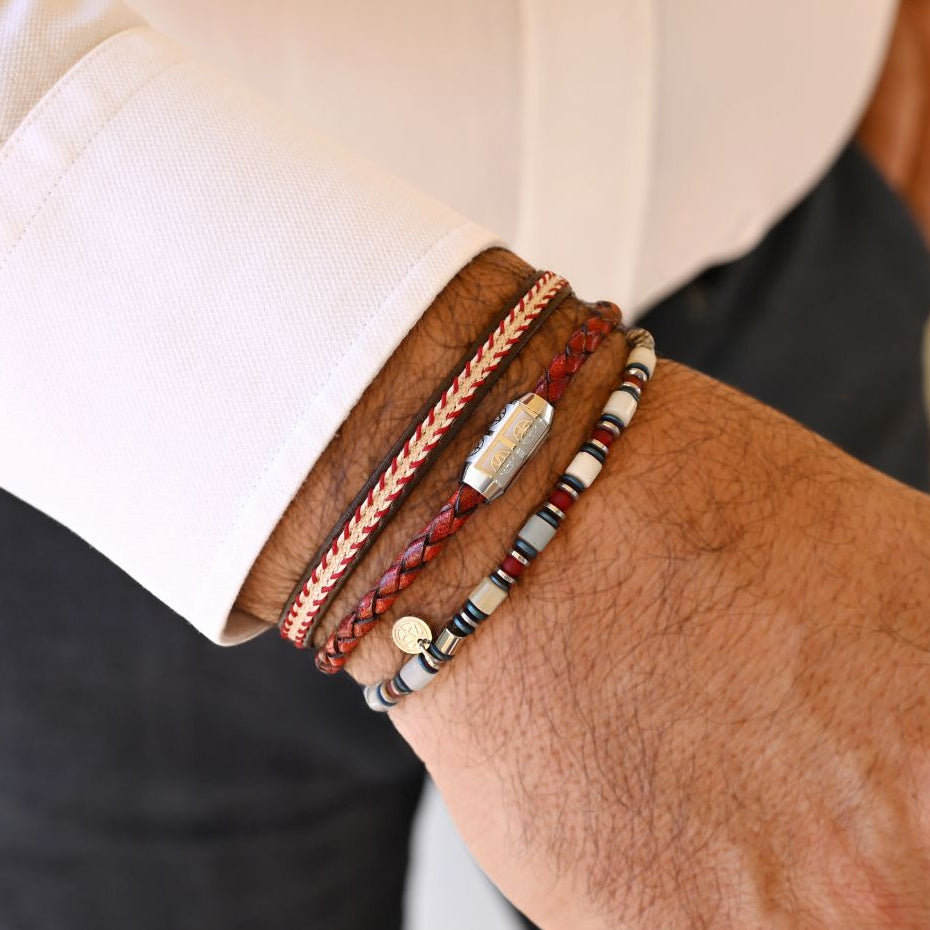 A close-up of a man's wrist wearing a stack of four distinct bracelets, including braided leather, beaded cord, and silver-toned metal bands, paired with a white dress shirt and dark trousers.